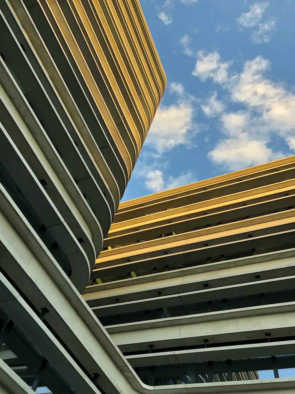 Modern multi-storey building with curved balconies viewed from below against a blue sky with scattered clouds.
