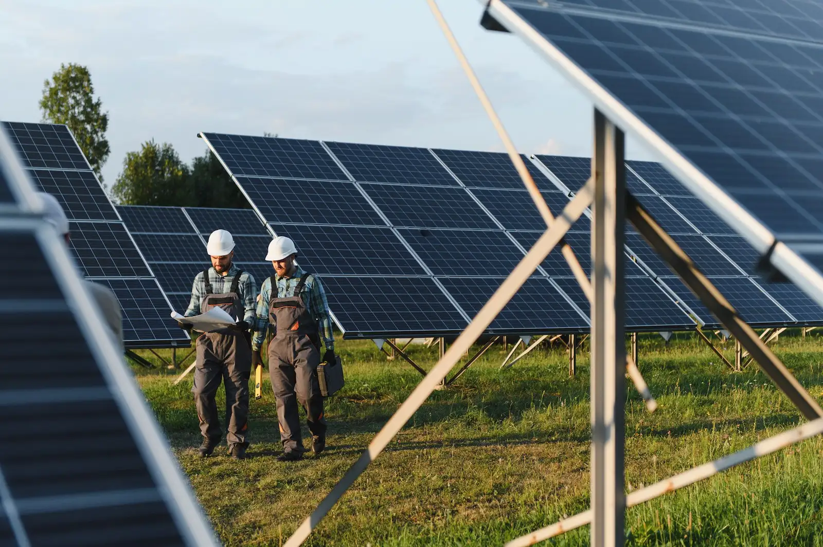 Two solar technicians wearing hard hats inspect plans while walking between rows of ground-mounted solar panels in a large solar farm on a sunny day.